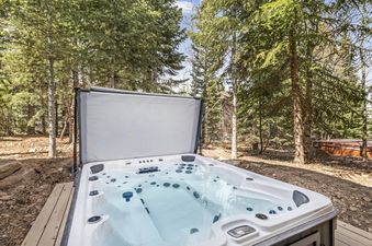A sparkling hot tub with its cover open, set on a wooden deck amidst a lush pine forest, with another hot tub visible in the background.