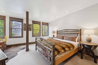 The bedroom at Woodrun Place 20 features a king-sized bed with a brown faux fur throw and a wood frame, a bedside table with a lamp, and windows with views of the trees.