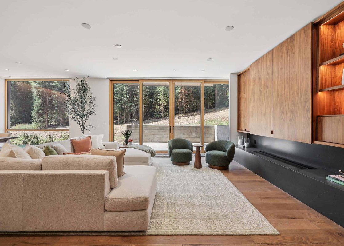 The living area at Founders Place 1403 showcases a modern design with plush seating, full-height windows offering forest views, and a stylish fireplace integrated into wooden paneling.