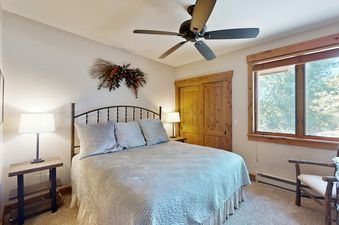 A bedroom with a queen-sized bed, a ceiling fan, and a window with a view of trees.
