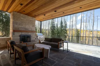 A luxurious outdoor patio at The Summit with a stone fireplace, BBQ grill, and modern lounge seating, overlooking a vibrant forest with aspen trees and mountains.