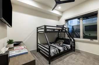 A modern bunk room at Frostwood Links features a black metal bunk bed with geometric patterned bedding, a wall-mounted TV, and a large window overlooking an outdoor area, creating a comfortable and functional sleeping space.