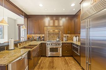 A fully equipped gourmet kitchen in Guardsman Overlook, featuring rich wooden cabinetry, stainless steel appliances, a hammered copper sink, granite countertops, and an earth-toned tiled backsplash.