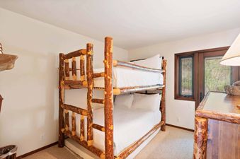 The rustic log-style bunk beds with white bedding are set against a light-colored wall with a window offering a view of lush greenery outside.
