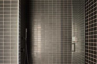 A modern shower stall featuring dark grey rectangular tiles with light grout, a black ceiling-mounted rain showerhead, a handheld sprayer, a glass enclosure, and a small wooden stool on the tiled floor.