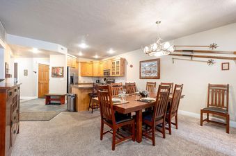 The dining area of Town Pointe A202 features a large, dark wood dining table set for six, illuminated by an antler chandelier, with a kitchen visible in the background.