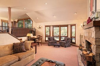 A spacious living area in Woodrun V Residence 5 features plush armchairs, a rustic stone fireplace, and large windows offering mountain views.