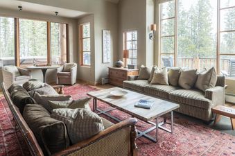 A cozy, rustic living room with plush sofas, a wooden coffee table on a red patterned rug, and large windows showcasing a snowy forest view.