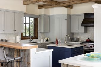 A modern rustic kitchen featuring light grey cabinets, exposed wood beams, and a navy blue island.