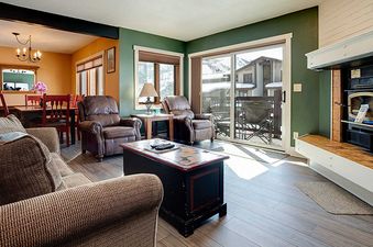 A cozy living room with a fireplace, brown sofas, and recliners, opening onto a balcony with snowy mountain views, adjoined by a dining area with a wooden table and red chairs.