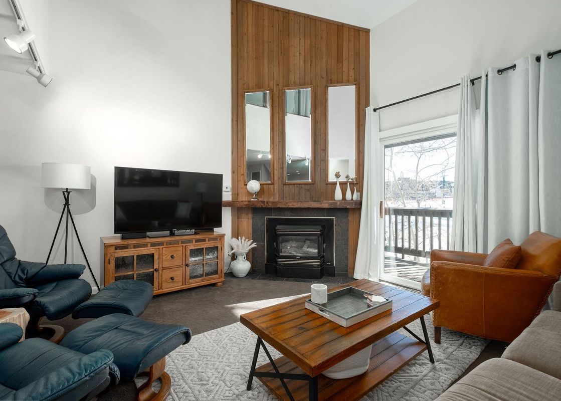 A modern and inviting living room at Snow Flower 129 Park City, featuring a large flat-screen TV, comfortable seating, a stylish coffee table, and a cozy fireplace with a wooden accent wall, and a balcony with an exterior view of a winter landscape.
