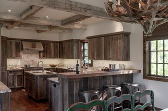 A rustic kitchen at Four Pines 09 with dark wood cabinetry, granite countertops, and modern stainless steel appliances, featuring a bar area with wine and stools and a large window with an outdoor view.