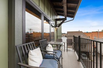 A private balcony at Baker Loft 301 with outdoor seating, a small dining table, and a black metal railing, overlooking a street view.