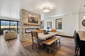 The dining area at Moonlight Ridge features a long wooden table with a matching bench and chairs, positioned in front of a stone fireplace, with a view of the mountains through large windows.