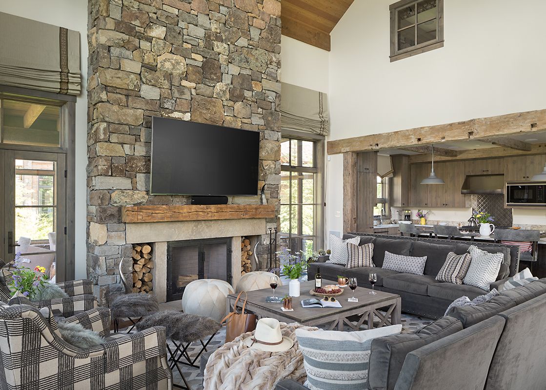 A spacious living room with a large stone fireplace, a wall-mounted TV, comfortable gray sectional sofa, and plaid armchairs, openly connected to a modern kitchen.