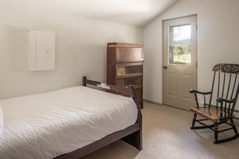 A serene bedroom at Glenn House featuring a single bed with white linens, a wooden bureau, a classic rocking chair, and a window with an exterior view.