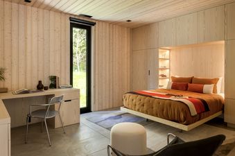 A modern bedroom with light wooden walls and ceiling, concrete floor, an orange bed with striped blanket, a wooden desk with a metal chair, and a tall window looking out to green trees.