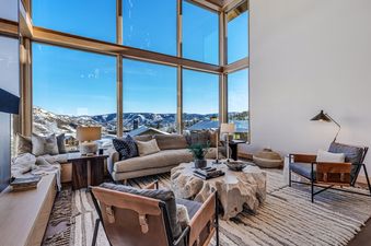 A luxurious living room at Havens Haus features floor-to-ceiling windows with panoramic views of snow-covered mountains, a modern sofa, and unique wooden coffee table.
