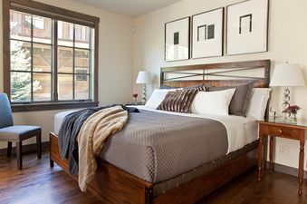 A king-sized bed with dark gray bedding and faux fur pillows, a wooden nightstand with a lamp, and a large window with a view of wooden buildings outside.