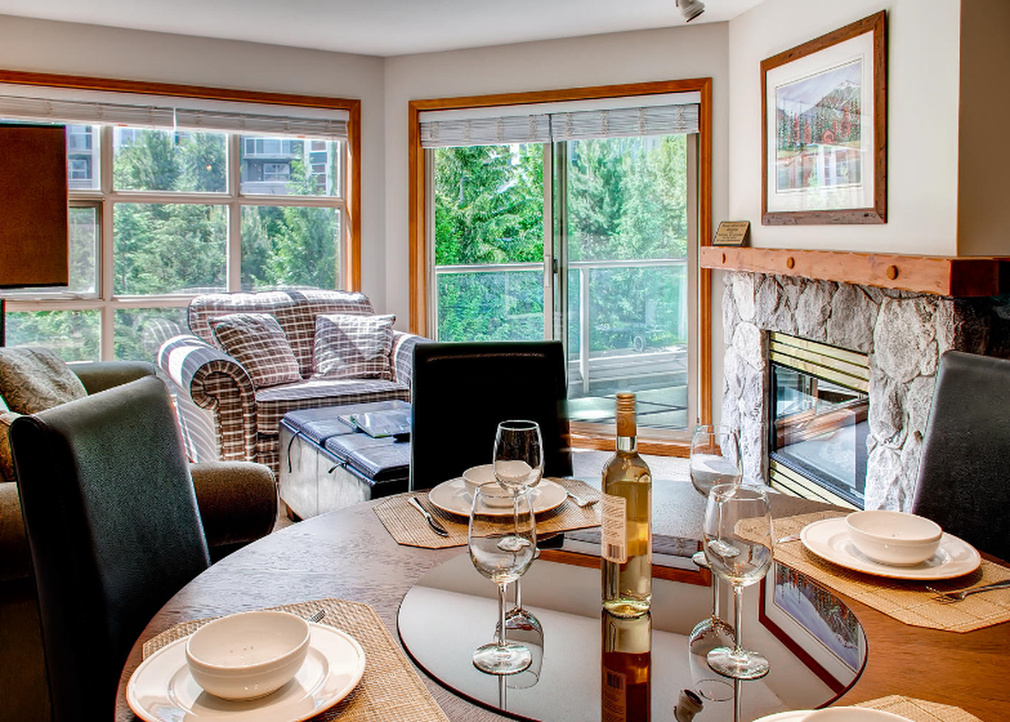 A cozy living area features a dining table set for guests, a comfortable armchair, and a fireplace, all bathed in natural light from large windows offering a view of lush greenery.
