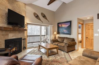 The image shows a cozy living room with a fireplace, comfortable seating, and a balcony view of a snowy landscape.