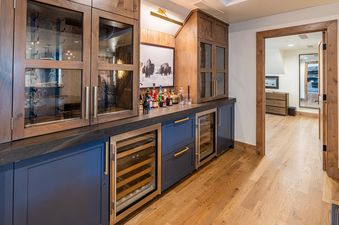 A sophisticated bar area in the Ritz Carlton West Wing, featuring dark wood cabinetry, glass display cases, a dark countertop with spirits, and integrated wine refrigerators, all set against warm wooden flooring.