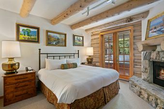 The bedroom in Pine Inn 09 features a king bed with a white duvet, decorative pillows, and a fireplace. The room is decorated with rustic log walls and wooden beams. There are windows on the back wall with views of the surrounding trees.