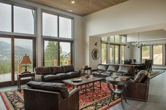 A spacious living room featuring leather sofas and armchairs arranged around a wooden coffee table on a vibrant red and black patterned rug, with large windows overlooking a scenic mountain landscape.