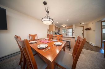 A spacious dining area with a wooden table set for six, featuring elegant place settings, and a large, decorative overhead light fixture. The adjacent kitchen is visible with stainless steel appliances and a breakfast bar.