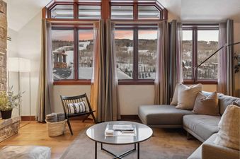 A living room with a large sectional sofa, a coffee table, and a rocking chair. The room has large windows with a view of a snowy mountain landscape.