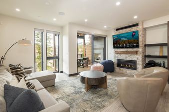 A modern and cozy living room featuring a large sectional sofa, a round coffee table, a fireplace set in stone, a flat-screen TV, and large windows looking out onto a wooded area.