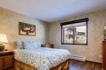 A cozy bedroom with a comfortable bed, bedside table with a lamp, a wooden cabinet, and a TV on a dresser, with a window providing natural light.