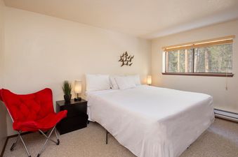 A cozy bedroom with a queen-sized bed, two nightstands, two lamps, a red chair, and a window offering a view of a forest.