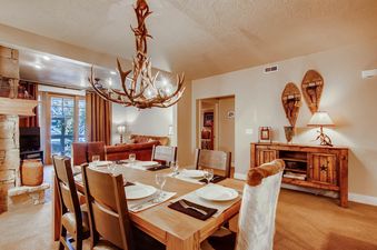 The dining area at Town Pointe B203 features a wooden table set for six, illuminated by an antler chandelier, with a rustic-chic design that blends seamlessly with the adjacent cozy living space.