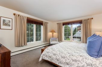 The bedroom in Chateau Christian 2 Bedroom Platinum Unit 210 features a queen-sized bed, two large windows with views of the mountain, a patterned armchair with a reading lamp, and light tan curtains.