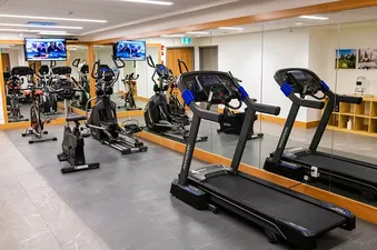 A bright and modern fitness center with treadmills, elliptical trainers, and stationary bikes, all facing mirrors and wall-mounted televisions.