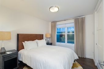 A bedroom with a queen bed, white linens, and a brown leather headboard. There is a window with beige curtains, a wood floor, and a nightstand with a lamp.