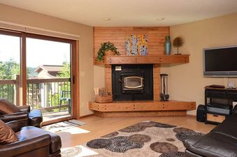 A cozy lounge area with leather furniture, a fireplace, and a TV, featuring large sliding glass doors that open to a balcony with a view of trees and houses.