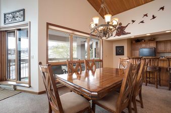 A spacious dining area with a large wooden table and chairs, a chandelier, and a bar area, offering views through a large window and sliding glass door.