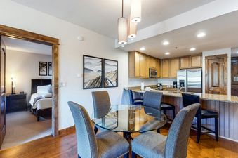 A dining area with a glass table and chairs, a view into a bedroom, and a kitchen in the background.