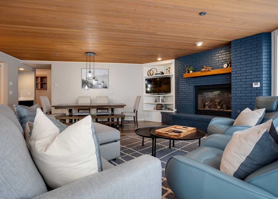 A spacious and modern living room and dining area in Chateau Chamonix CX113 with a blue brick fireplace, a grey sectional sofa, blue armchairs, and a large wooden dining table.