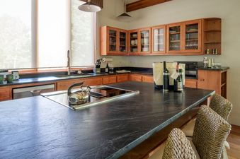A modern kitchen with wooden cabinets, dark countertops, an island stovetop, and bar stools, featuring a large window with an exterior view.