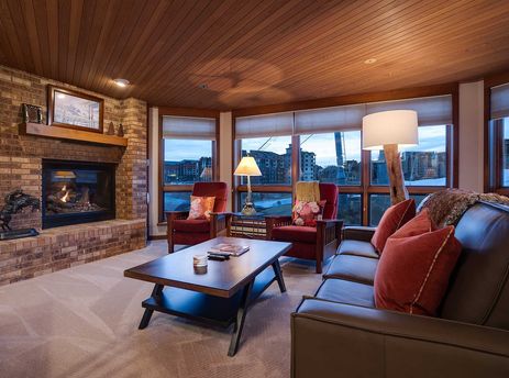 The Chateau Chamonix CX123 living room showcases a warm fireplace, a brown leather sofa with red pillows, and two accent chairs by large windows offering scenic views.