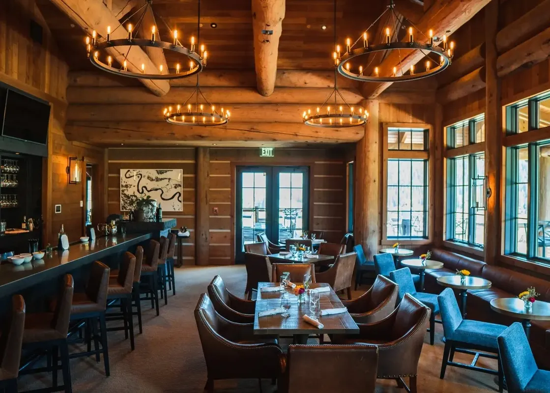 A rustic-chic dining area and bar within The Sylvan Lodge One Bedroom Penthouse, featuring wooden interiors, elegant chandeliers, and set tables with views of the outdoors.