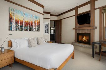 A spacious bedroom at Rocky Road Hideaway features a king-sized bed with white linens, wooden nightstands, and a vibrant painting of birch trees above the headboard. A fireplace with a TV mounted above it is on the adjacent wall.