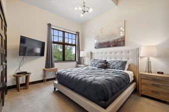 The bedroom at 2nd Street Lofts 209 features a comfortable bed with a tufted headboard, dark grey bedding, a wall-mounted TV, and a large window with green outdoor views.