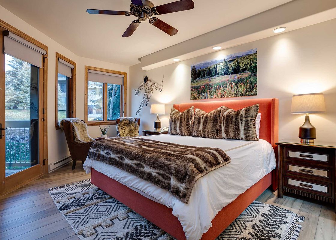 The bedroom at Chateau Chamonix CX322 features a large bed with a vibrant orange headboard, faux fur bedding, two nightstands with lamps, a cozy armchair, and large windows offering an outdoor view.