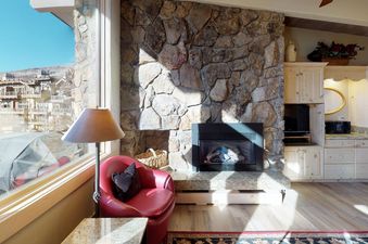 The living room of Manor Vail 364 features a fireplace and a view of the surrounding mountains from the window. The fireplace is lit, creating a warm and inviting atmosphere.