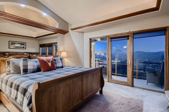 The bedroom at Moose on the Loose features a large bed with checkered bedding, a red throw pillow with a deer motif, and direct access to a balcony with scenic mountain views through glass doors.