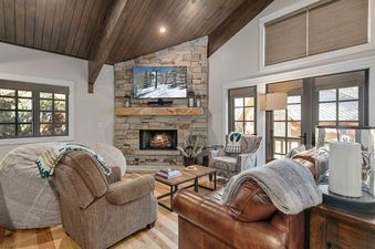 A cozy living room with a vaulted wooden ceiling, stone fireplace with a TV showing a snowy mountain scene, and comfortable seating with throws.